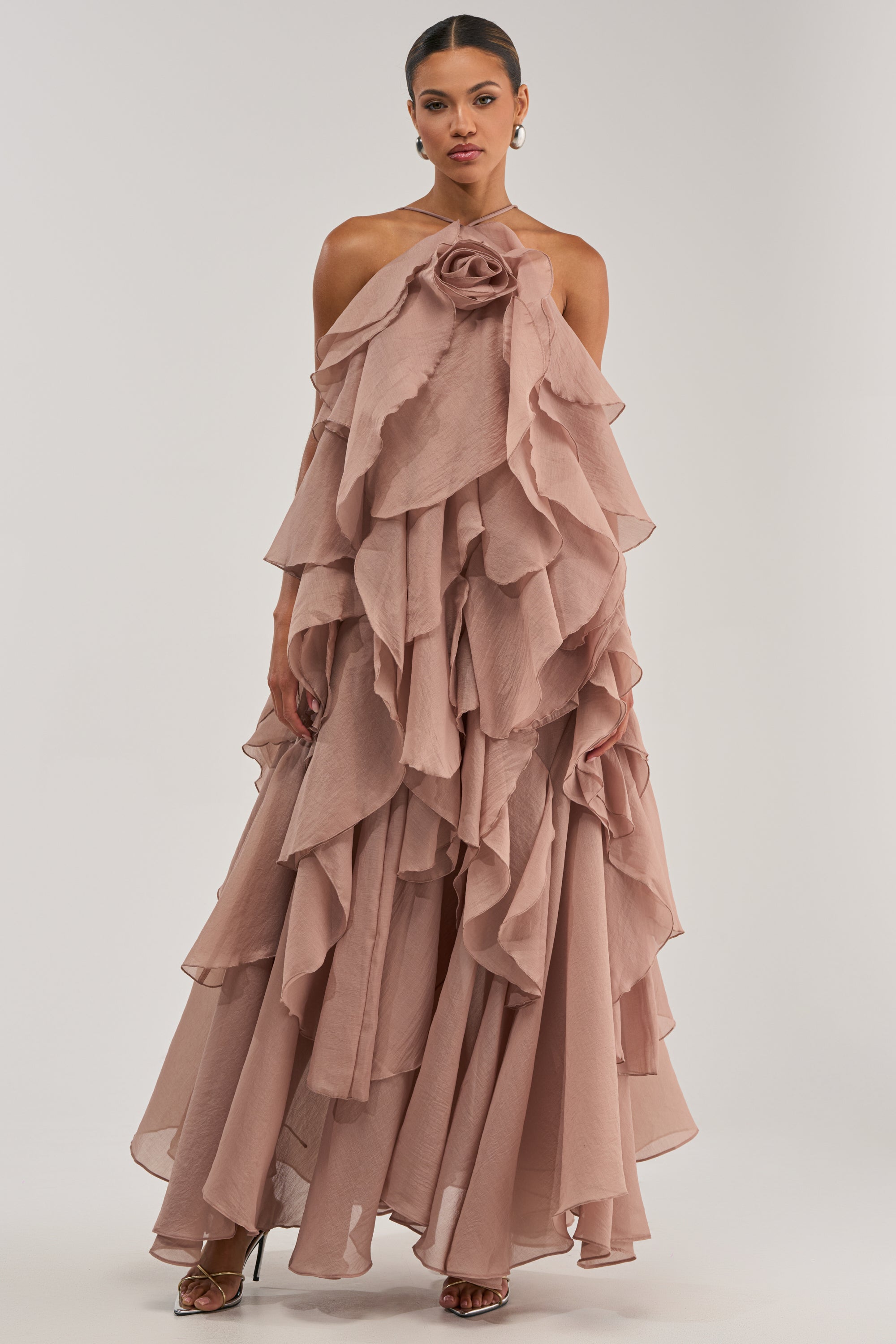 A woman wears the GOLDEN HOUR FLORAL CHIFFON MAXI DRESS IN MAUVE, featuring flowing, tiered ruffles, textured chiffon layers, and a large rose detail at the neckline, set against a plain white background.