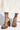 A close-up shows someone wearing AKIRA KNAPP BROWN CORK PLATFORM SANDALS with chunky heels and brown leather straps. The tall, black suede-textile block heel pairs beautifully with their light-colored, flowing skirt.