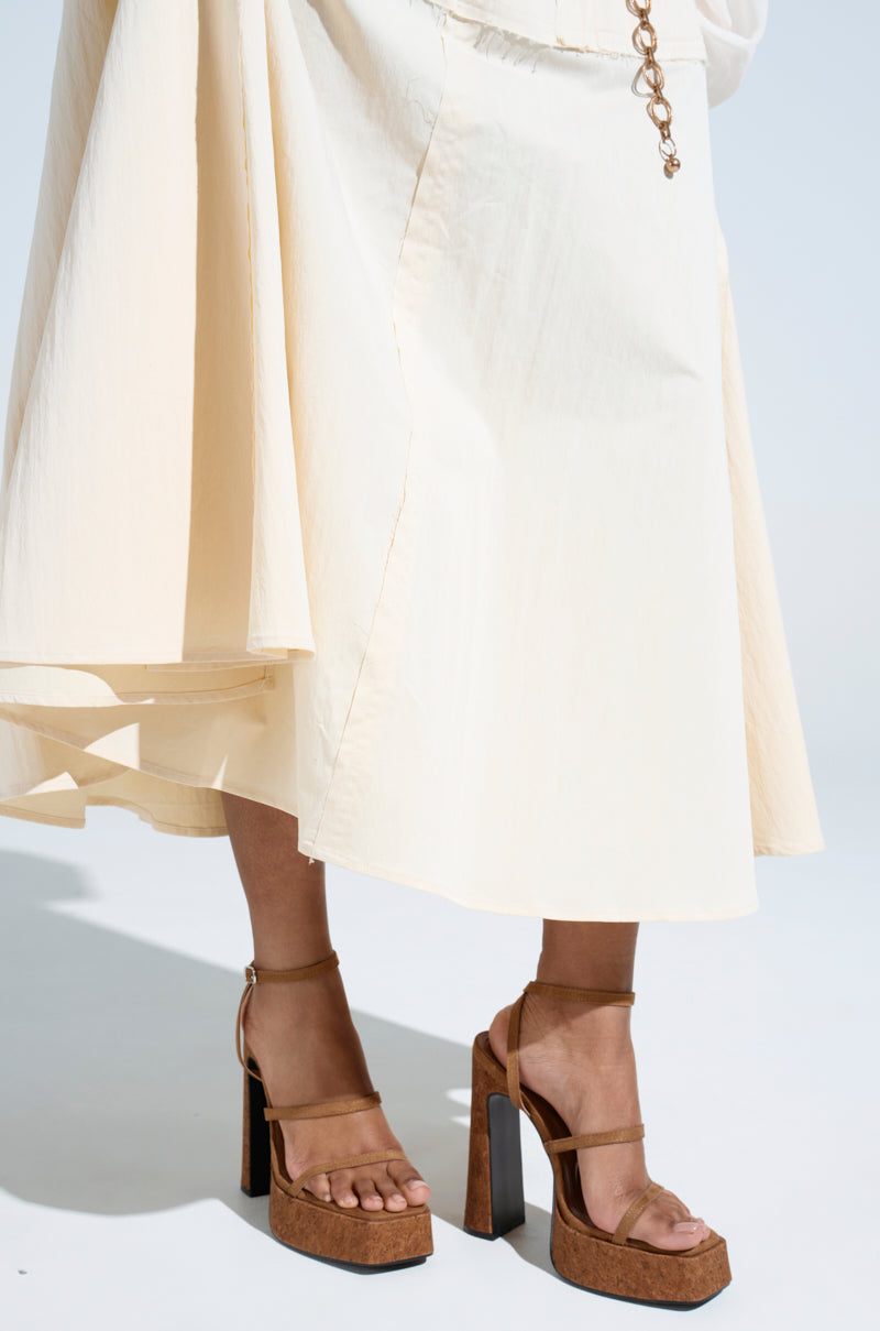 A person in a cream dress stands on a light background, pairing it with the AKIRA KNAPP BROWN CORK PLATFORM SANDAL, which has ankle straps for an earthy vibe. The dress's flowing fabric accentuates the platform sandal's bold style.