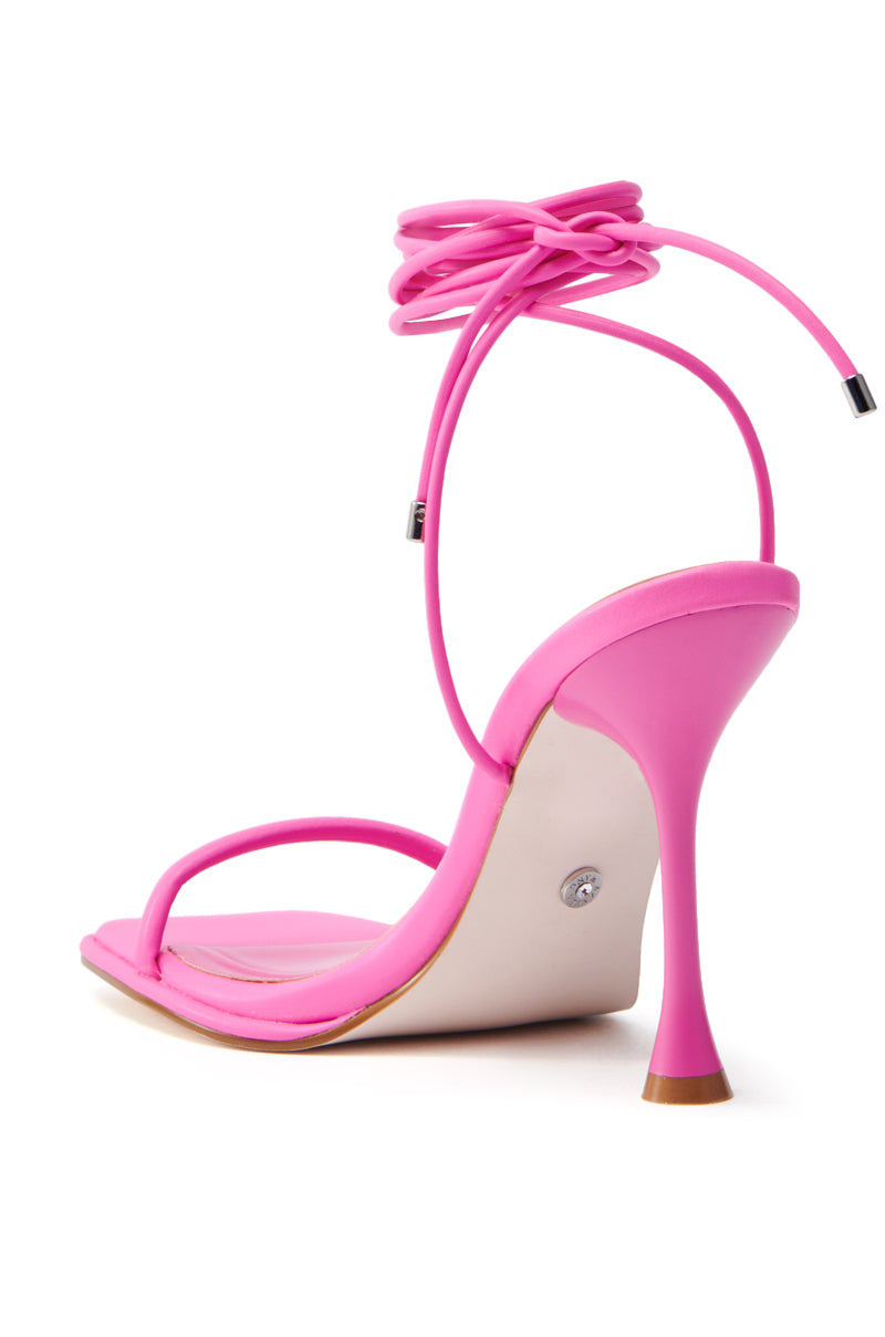 The AZALEA WANG KOHINOOR PINK BOOTIE STILETTO is a vibrant pink sandal made from vegan leather, featuring a slender stiletto heel and wrap-around ankle strap, with an elegant minimalist design and smooth shiny finish atop a beige sole.