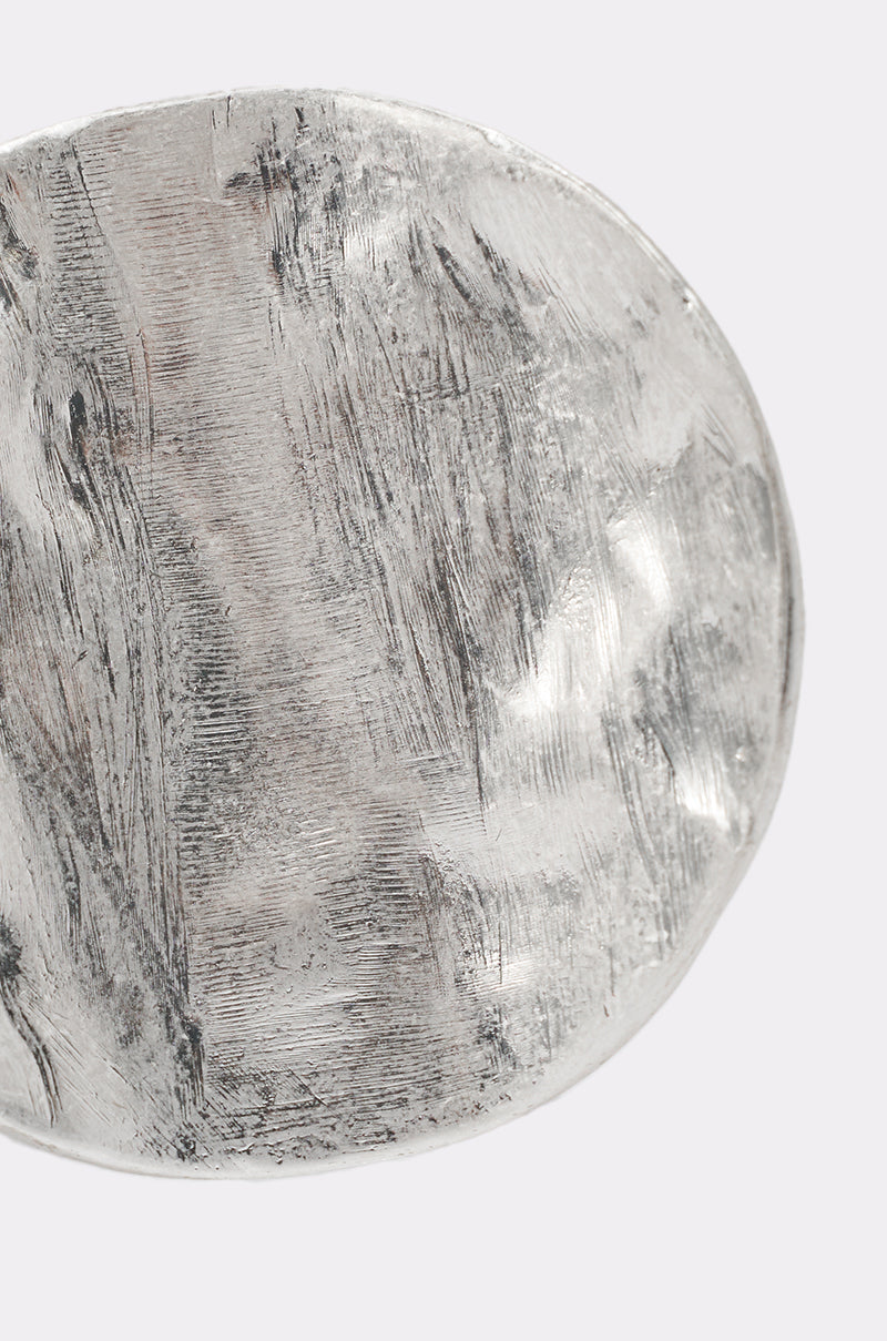 Close-up of the MOLTEN DREAMS RING, a round, textured silver-tone statement piece featuring visible brush strokes and an irregular edge, set against a light gray background. 4