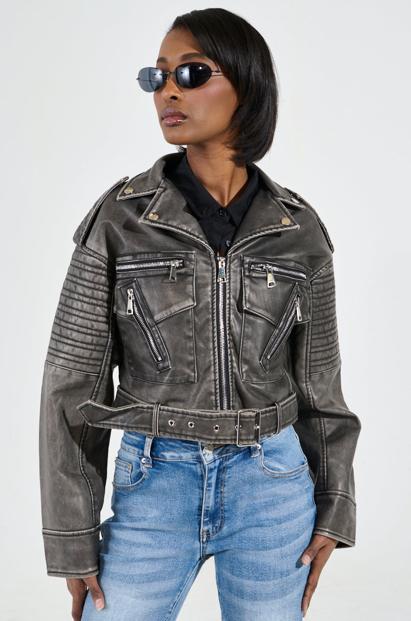 A person styled in a black faux leather jacket featuring quilted sleeves and zippered pockets is paired with SAY MY NAME DISTRESSED MOTO jeans and sunglasses. The ensemble exudes a bold, edgy vibe, highlighted by metallic hardware, as the individual strikes a confident pose against a plain background.