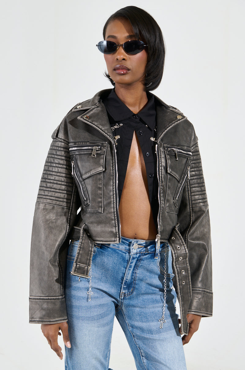 A person wearing the SAY MY NAME DISTRESSED MOTO jacket, with its metallic hardware, stands confidently. Their dark faux leather attire is elegantly draped over a partially open black shirt and blue jeans. Sporting short hair and dark sunglasses, they pose against a plain background.
