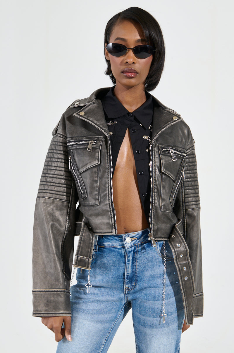 A person wearing the SAY MY NAME DISTRESSED MOTO, a black faux leather jacket featuring metallic hardware, paired with a black open-front shirt and blue jeans, stands against a plain background. They have short hair and dark sunglasses, exuding confidence with hands in pockets.