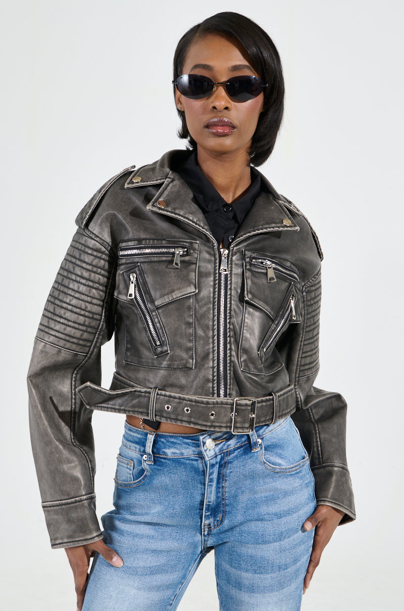 A person clad in the SAY MY NAME DISTRESSED MOTO jacket featuring metallic hardware, paired with blue jeans and black sunglasses. They strike a confident pose with their hands in pockets against a plain light background.