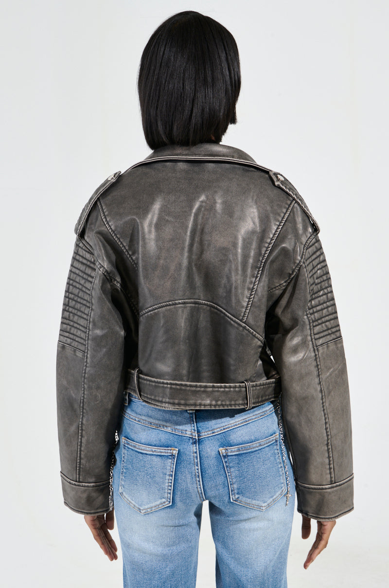 Someone is wearing the SAY MY NAME DISTRESSED MOTO jacket, which has a stylish appearance from the back with its quilted sleeves. The dark gray faux leather of the jacket showcases metallic hardware, and it is paired with light blue jeans against a plain white background.
