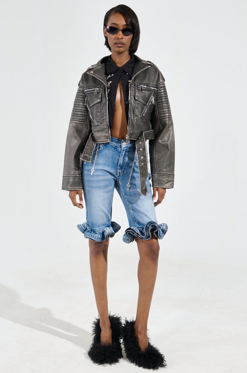 The fashion model looks chic in the SAY MY NAME DISTRESSED MOTO jacket, featuring sleek metallic hardware. She pairs it with a slightly unbuttoned black shirt, knee-length denim shorts with ruffled hems, and fluffy black slippers. Her outfit is flawlessly accessorized with sunglasses.