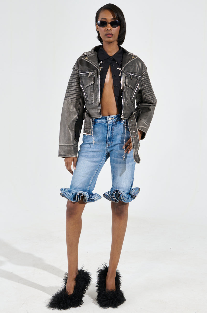 A stylish person in sunglasses wears the SAY MY NAME DISTRESSED MOTO black faux leather jacket over a black top, denim shorts with ruffled hems, and fluffy black shoes. They are standing against a plain white background.