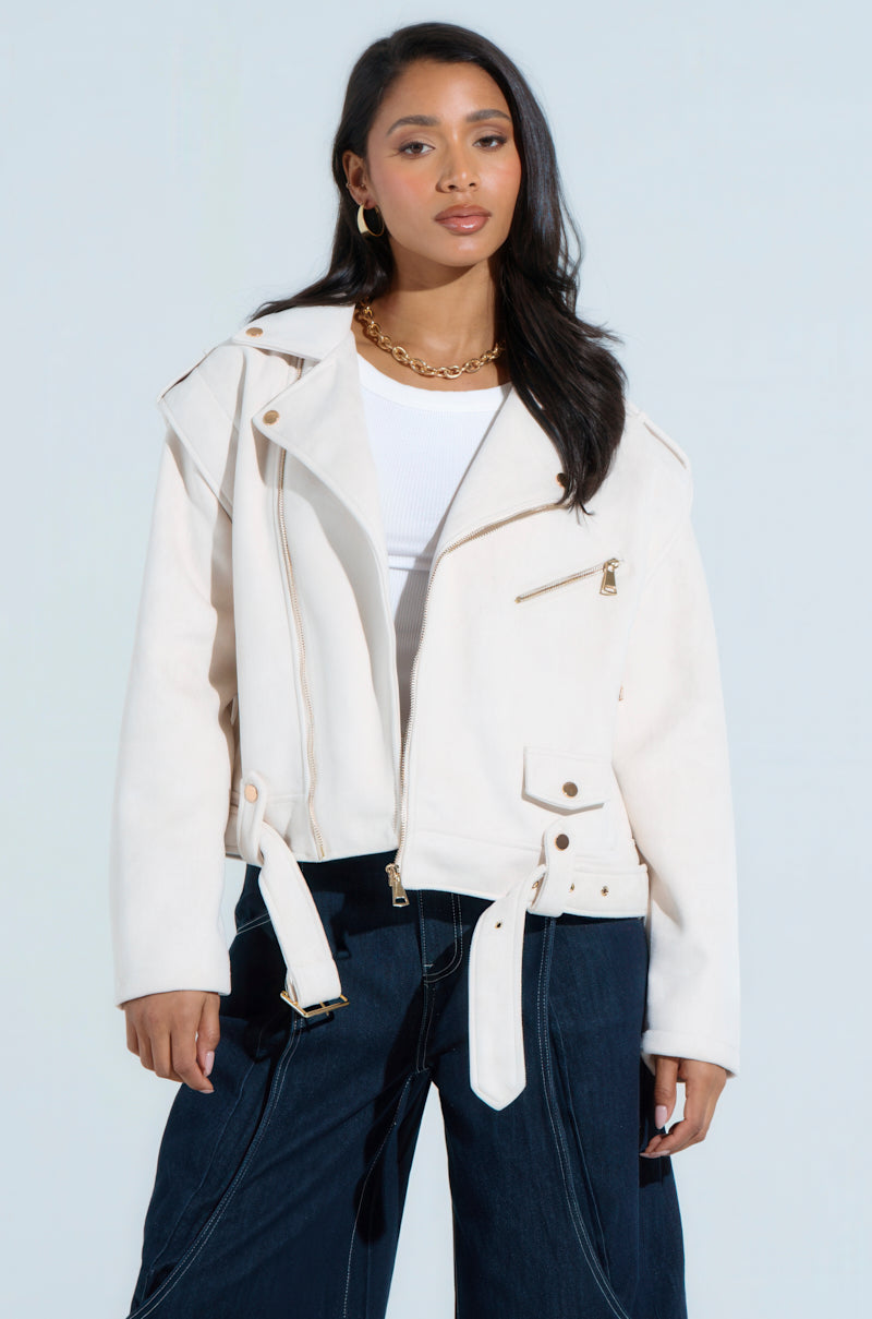 A person in chic outerwear poses against a light background, wearing the MONTE CARLO FAUX SUEDE MOTO jacket over a white top with dark wide-leg pants. They have long dark hair, accessorized with gold hoop earrings and a chain necklace, exuding effortless style in the boxy cropped silhouette.