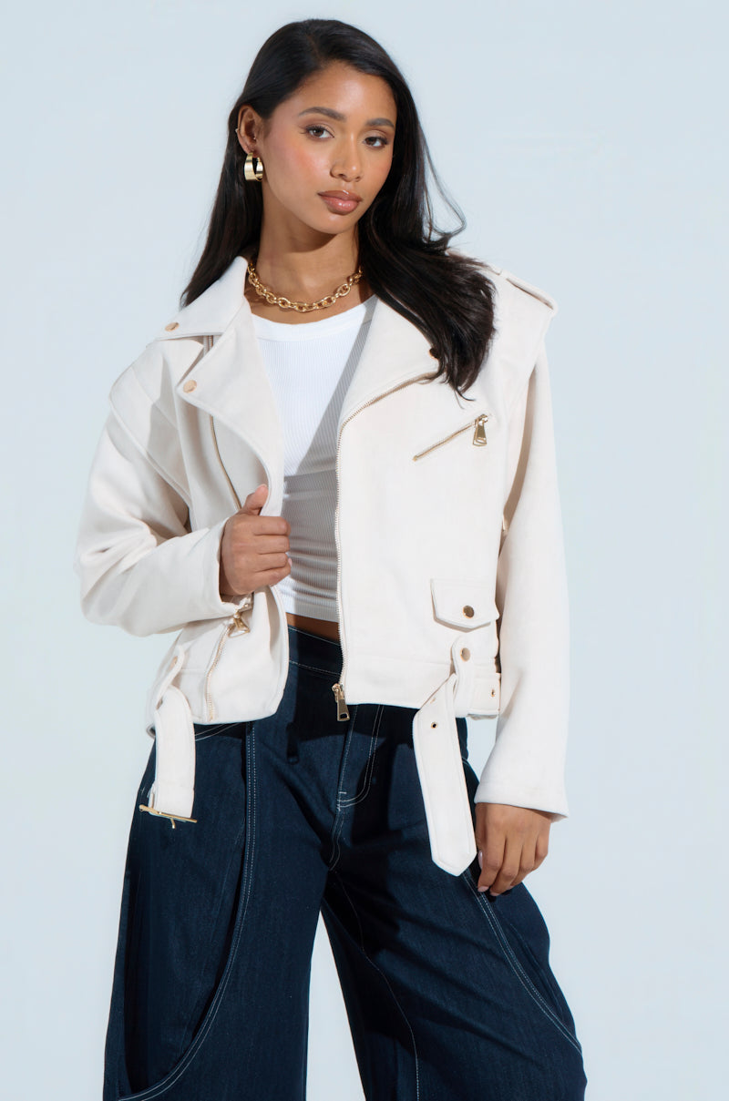 A woman confidently poses in a chic ensemble featuring the MONTE CARLO FAUX SUEDE MOTO jacket over a white top and dark wide-legged jeans. Her gold hoop earrings, necklace, and hand on her hip highlight the boxy cropped silhouette against a light background.