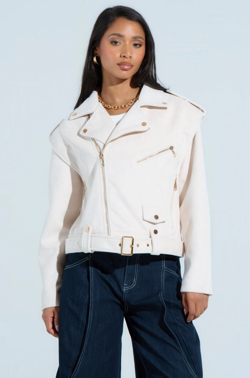 Wearing a chic MONTE CARLO FAUX SUEDE MOTO, a person pairs their look with a gold chain necklace and dark wide-leg jeans. With long, dark hair and a relaxed expression, they stand against a light gray background exuding effortless style.