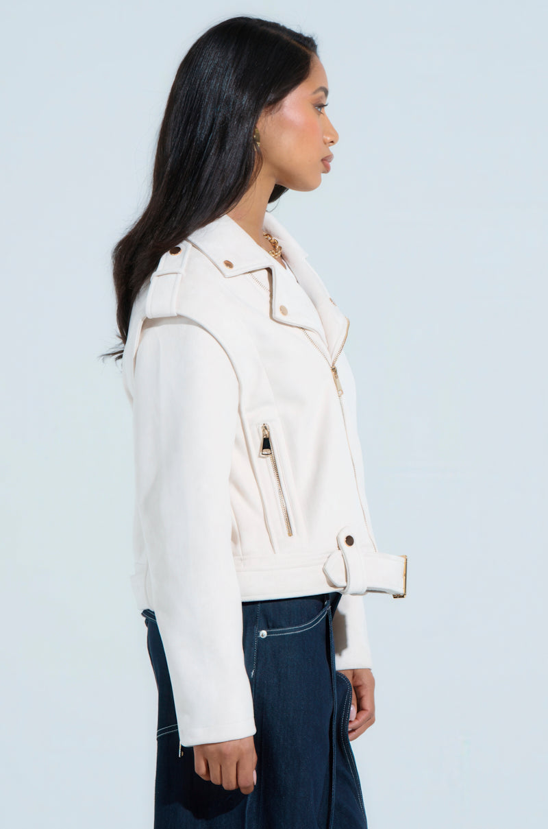 A person with long dark hair stands sideways, showcasing chic outerwear: the MONTE CARLO FAUX SUEDE MOTO jacket. This white belted design with gold zippers and buttons is paired with dark pants, and its boxy cropped silhouette adds a modern touch against the plain light background.