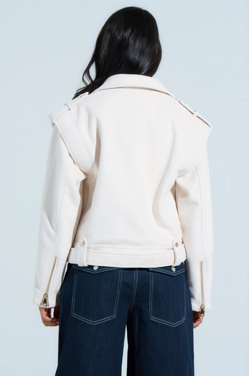 A person with long dark hair showcases the chic MONTE CARLO FAUX SUEDE MOTO jacket featuring broad shoulders, gold zippers on the sleeves, and a waist belt. Paired with dark wide-leg jeans, this ensemble exudes effortless style against a light background.