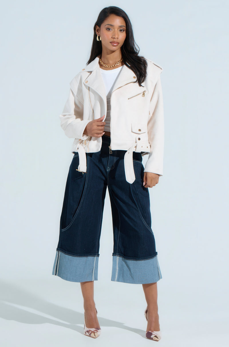A woman stands confidently, flaunting a MONTE CARLO FAUX SUEDE MOTO jacket in white over a matching top. She completes her chic look with wide-legged dark denim jeans with light cuffs, gold hoop earrings, a chain necklace, and snakeskin high heels.
