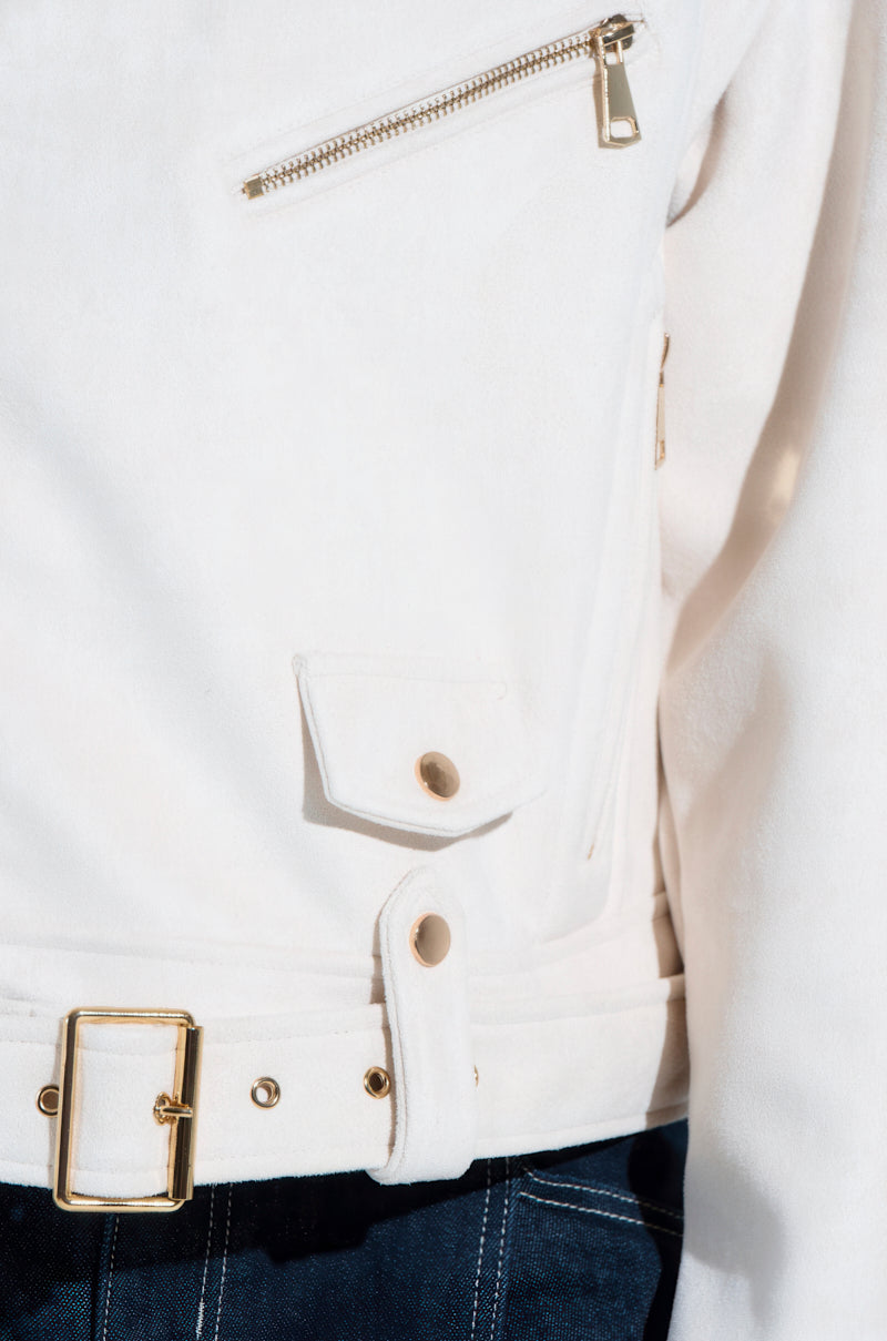 A close-up of the MONTE CARLO FAUX SUEDE MOTO: a white jacket with gold accents like a zippered pocket, flap pocket with button, and gold-buckle belt. The boxy cropped silhouette pairs perfectly with dark pants featuring visible stitching.