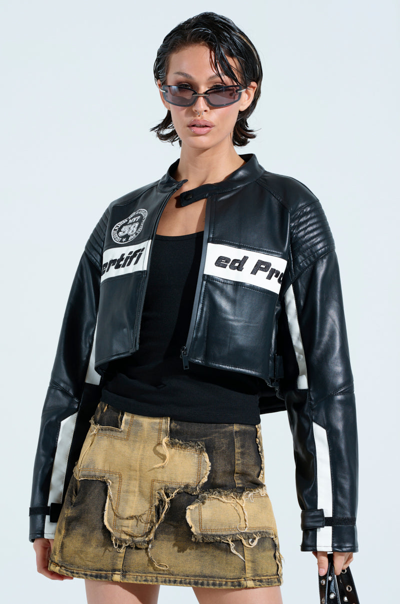 A model showcases the PRO LEAGUE BOXY MOTO jacket with white graphics, paired with a black top and distressed patchwork skirt. She completes her confident, edgy look with sleek sunglasses and a small bag.