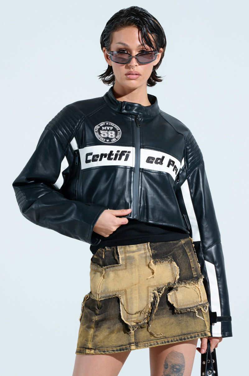 Wearing the PRO LEAGUE BOXY MOTO, a chic black jacket with white text, along with sunglasses and a distressed denim skirt with patches, a person with short dark hair and a bag stands against a plain light blue backdrop.