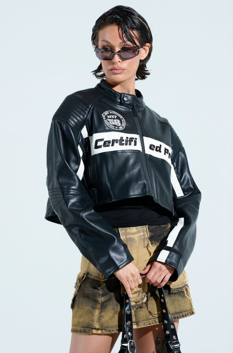 A person wearing the PRO LEAGUE BOXY MOTO jacket with white patches, sunglasses, and a tan denim skirt holds a strap against a light background.