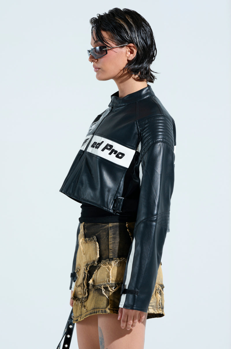 Wearing a PRO LEAGUE BOXY MOTO jacket with white text, a person in sunglasses and a black top pairs it with a distressed brown and black skirt. With short, dark slicked-back hair and light blue nail polish, they stand against a plain white background.