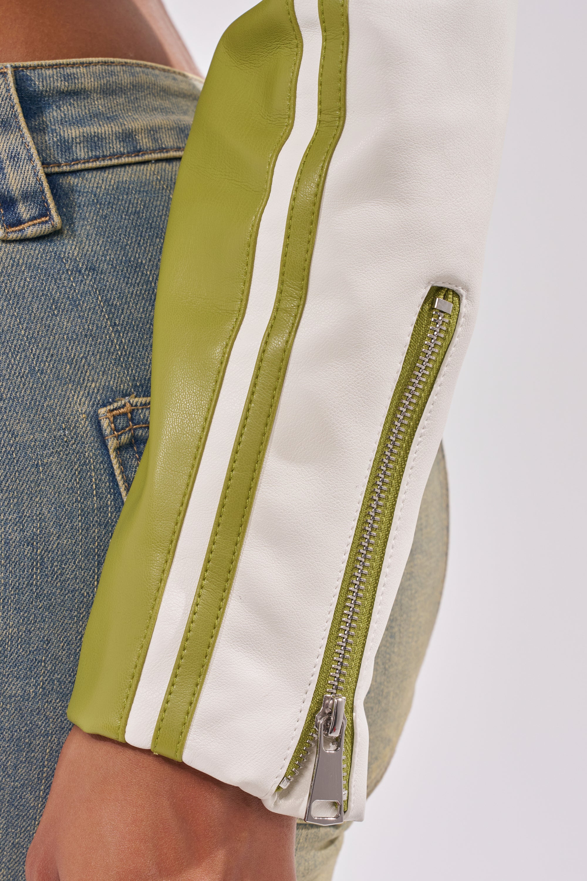 A close-up of a person wearing blue denim jeans and the WEST LOOP RACER MOTO IN GREEN with green and white sleeves, silver zipper, and white stripe detail—perfect for streetwear styling. 10