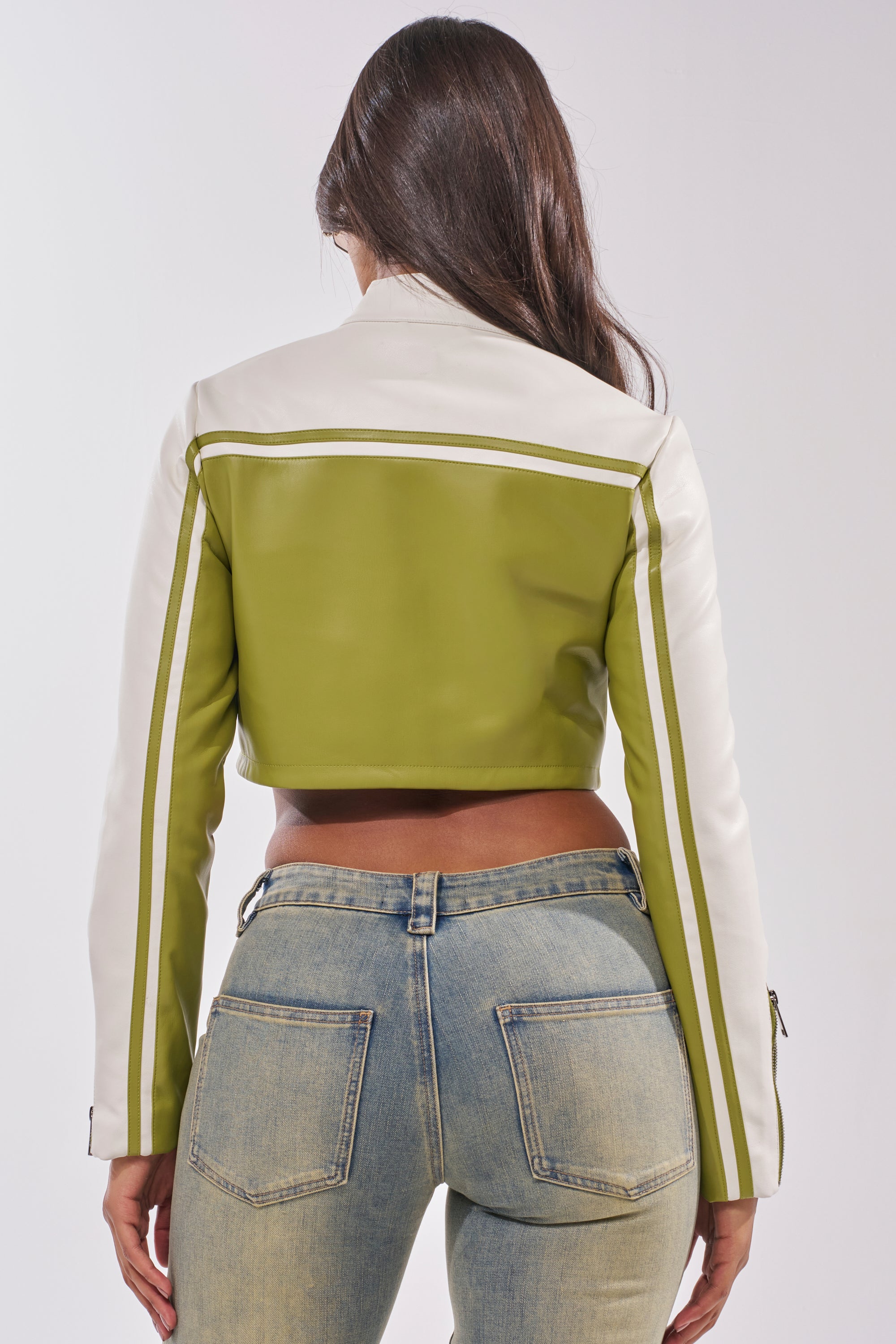 A woman faces away wearing the WEST LOOP RACER MOTO IN GREEN, a cropped faux leather jacket with long sleeves, paired with blue faded jeans. Her long brown hair is down and her hands rest at her sides against a plain background. 6