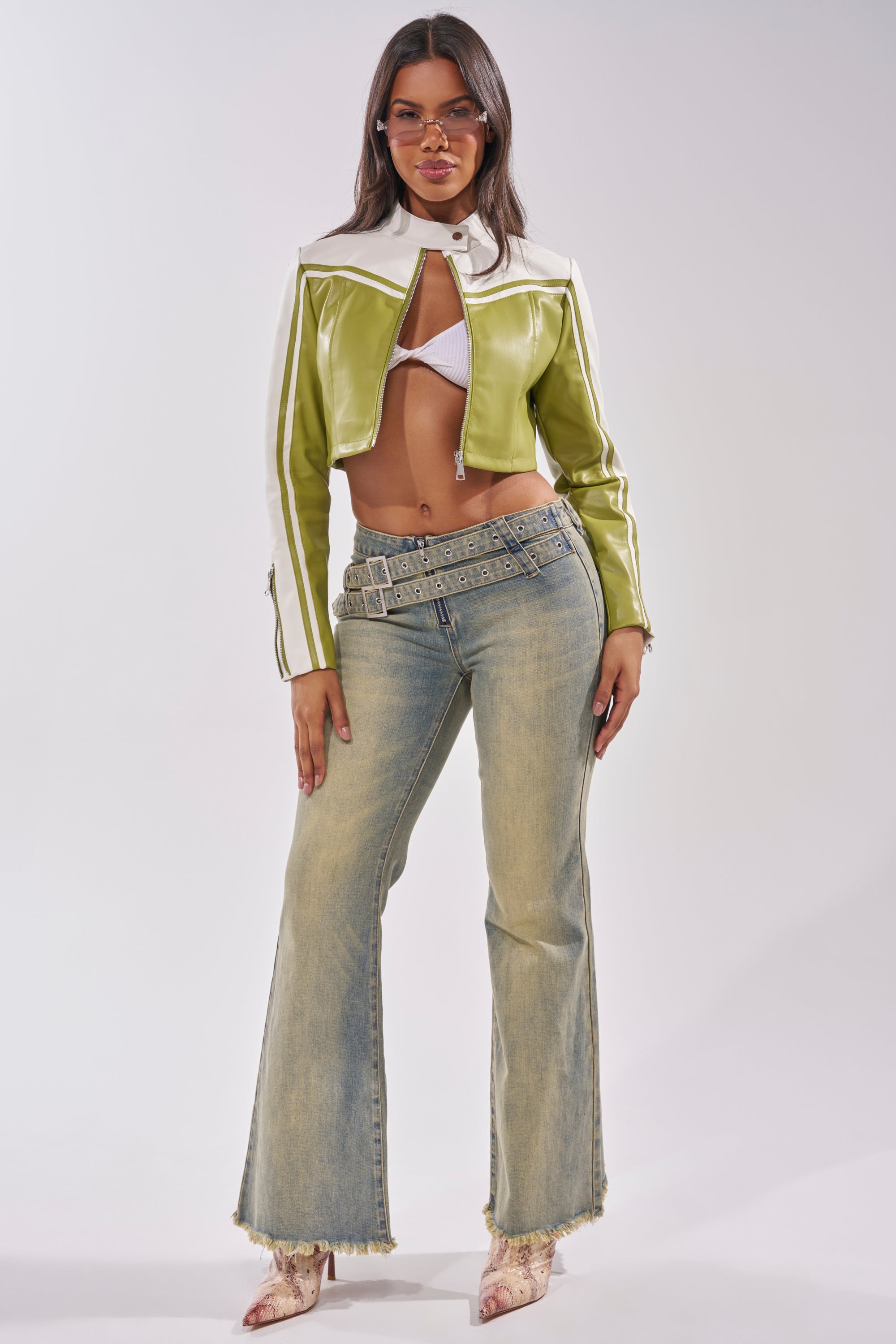 A woman poses in the WEST LOOP RACER MOTO IN GREEN, styled with a white bra top, high-waisted light-wash flared jeans, studded belt, and pointed-toe heels. She wears glasses and has long, straight hair. 7