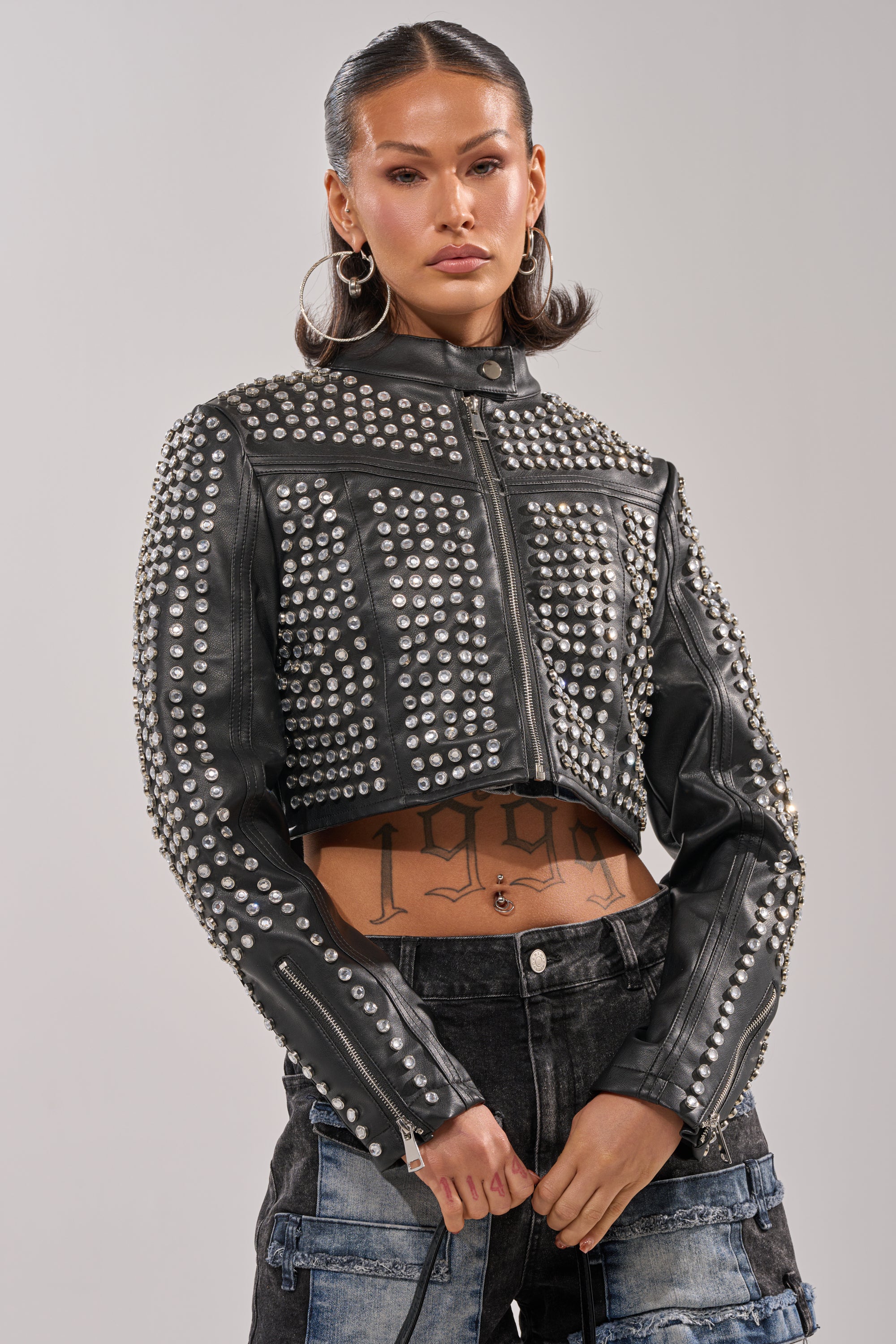 A woman stands confidently wearing the ASHTON RHINESTONE MOTO jacket with bedazzled studs, hoop earrings, dark denim pants, and visible midriff tattoos. Her slicked-back dark hair frames her direct gaze at the camera.