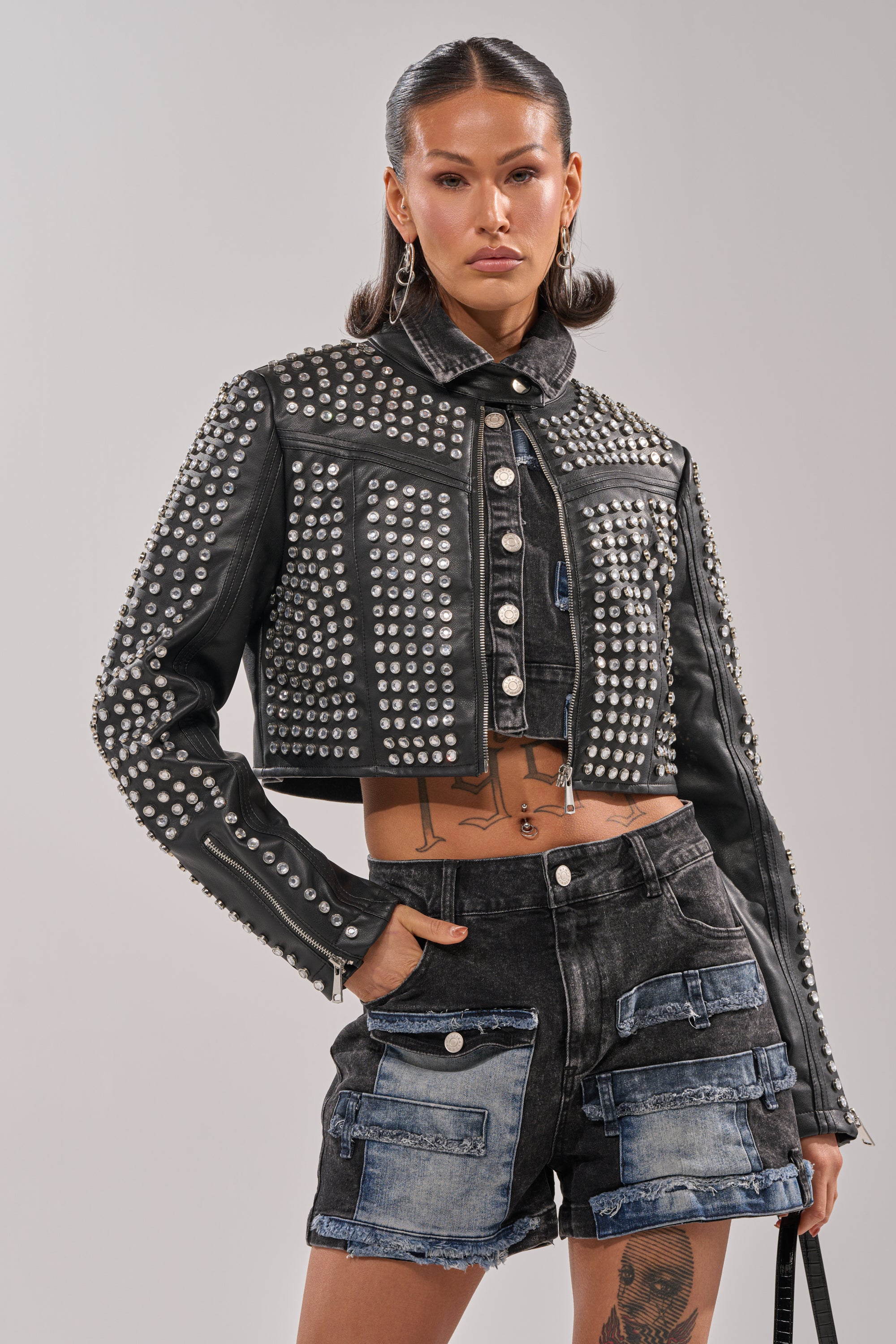Wearing the ASHTON RHINESTONE MOTO jacket, a person with slicked-back hair pairs it with black denim shorts featuring blue patchwork and multiple pockets, striking a confident pose against a plain background.