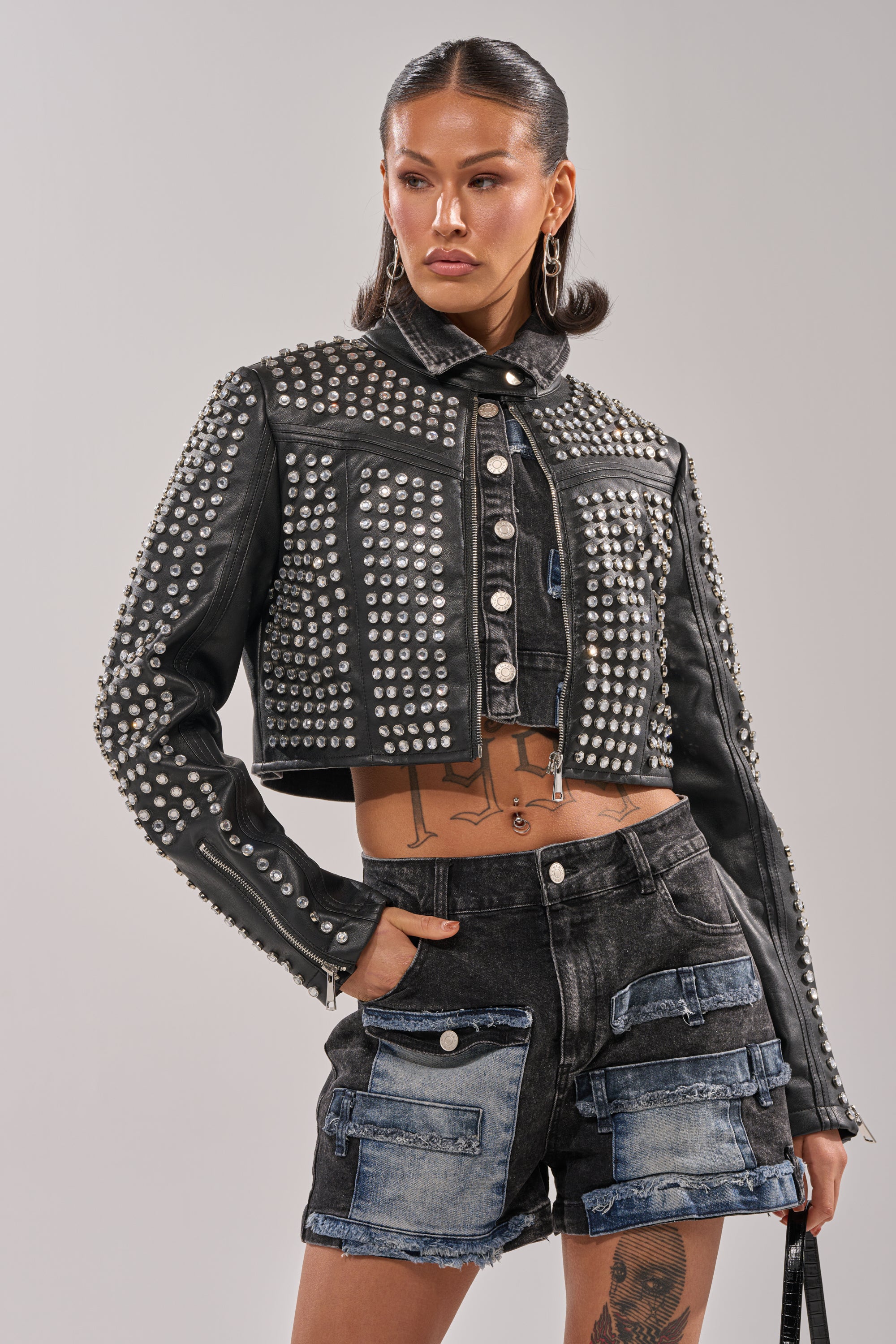 A woman poses confidently against a plain background, wearing the ASHTON RHINESTONE MOTO jacket with high-waisted black patchwork denim shorts, hoop earrings, and visible tattoos.