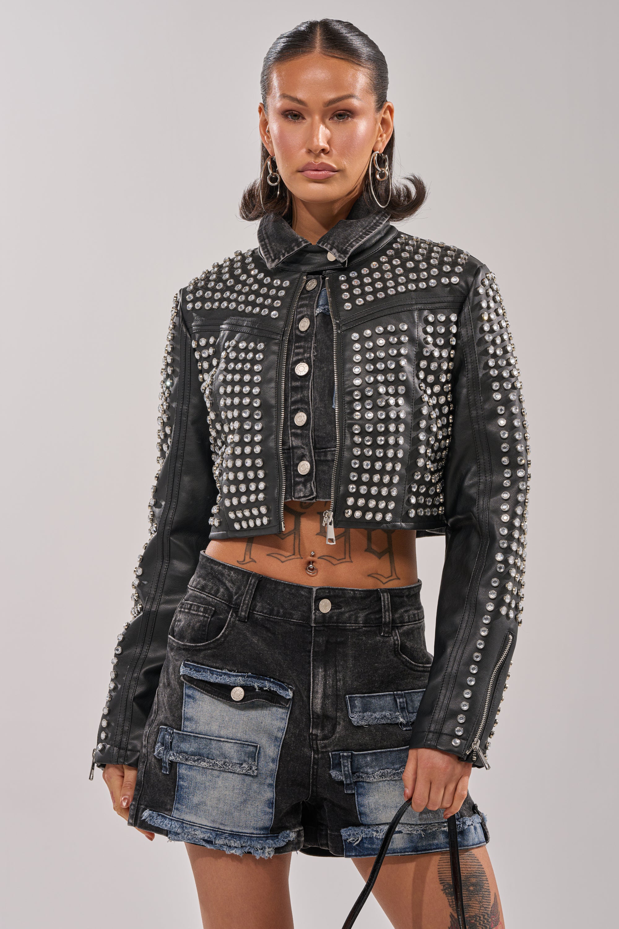 Wearing the ASHTON RHINESTONE MOTO jacket and black patchwork denim shorts with large front pockets, a woman shows off hoop earrings and tattoos while holding a bag against a plain light background.