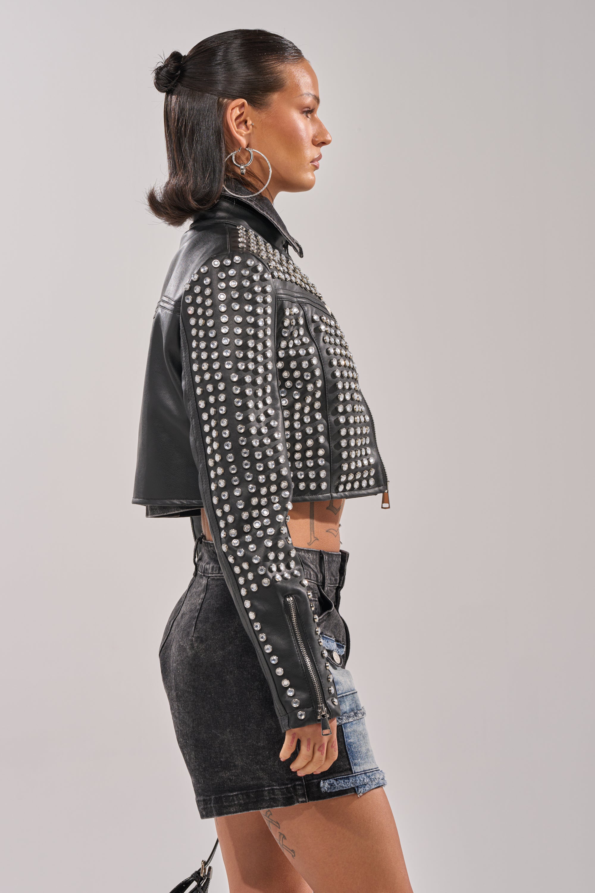 A woman with slicked-back dark hair stands in profile against a plain background, wearing the ASHTON RHINESTONE MOTO jacket, hoop earrings, and high-waisted black denim shorts.