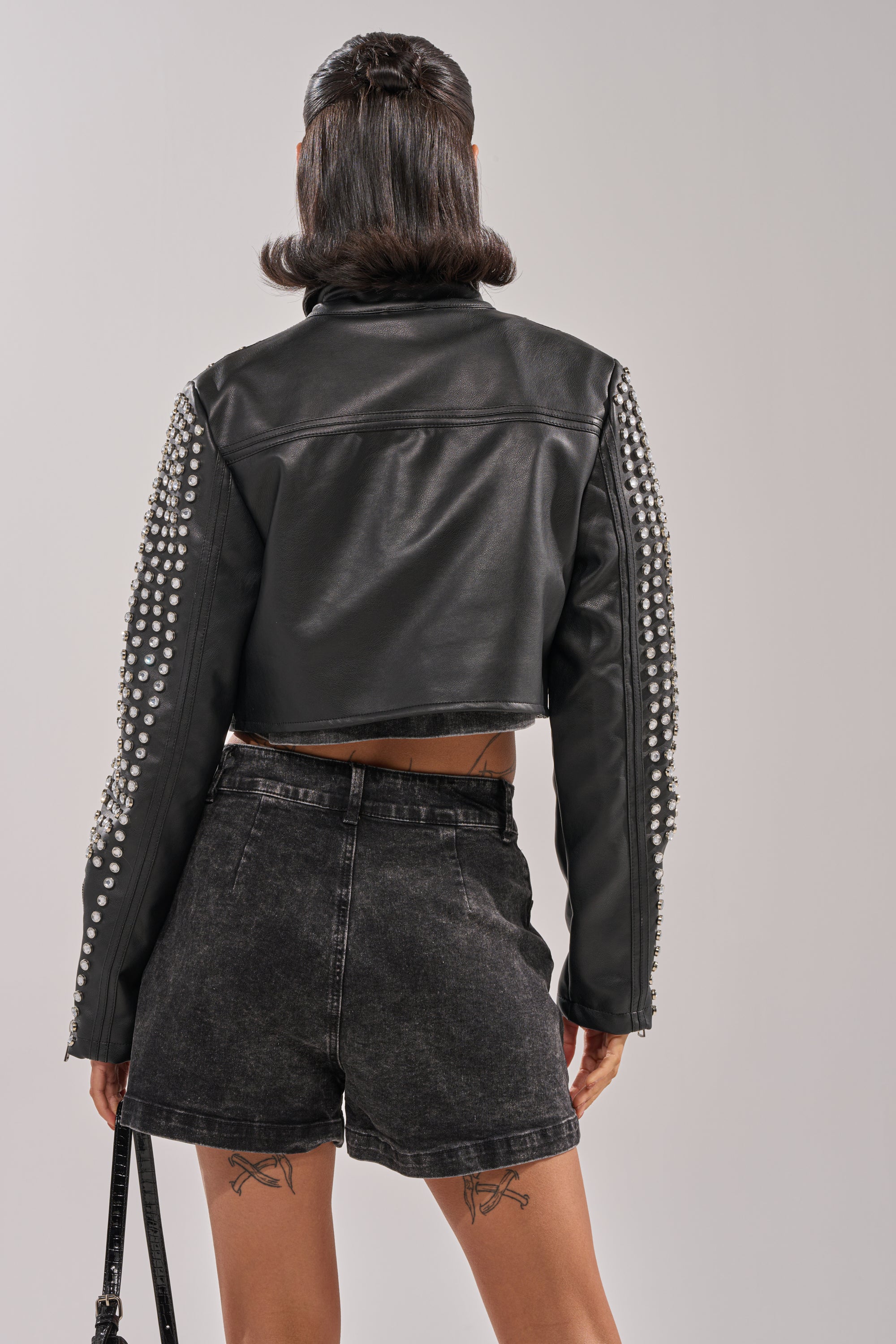 A person facing away wears the ASHTON RHINESTONE MOTO, a cropped black jacket with silver-studded sleeves, paired with high-waisted black denim shorts. They have shoulder-length dark hair and thigh tattoos.