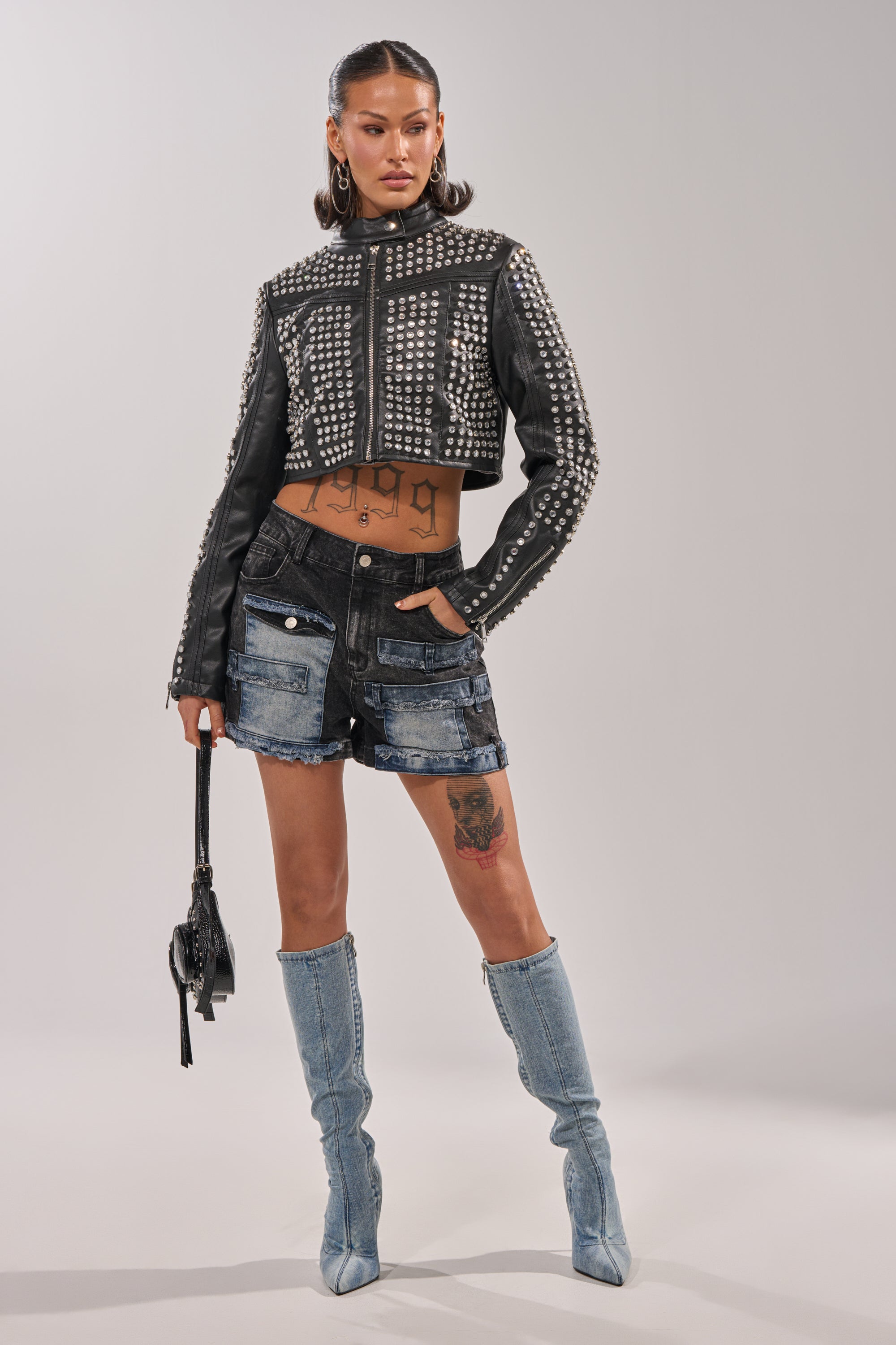 A woman poses confidently in the ASHTON RHINESTONE MOTO jacket, patched denim shorts, and light blue knee-high denim boots, holding a small black bag and displaying tattoos on her torso and left thigh.