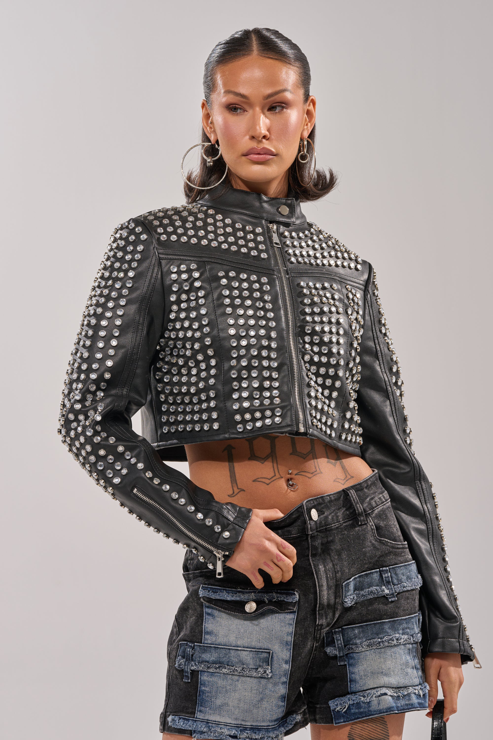Wearing the ASHTON RHINESTONE MOTO jacket over black denim shorts, a person with slicked-back hair and visible torso tattoos stands against a plain background.
