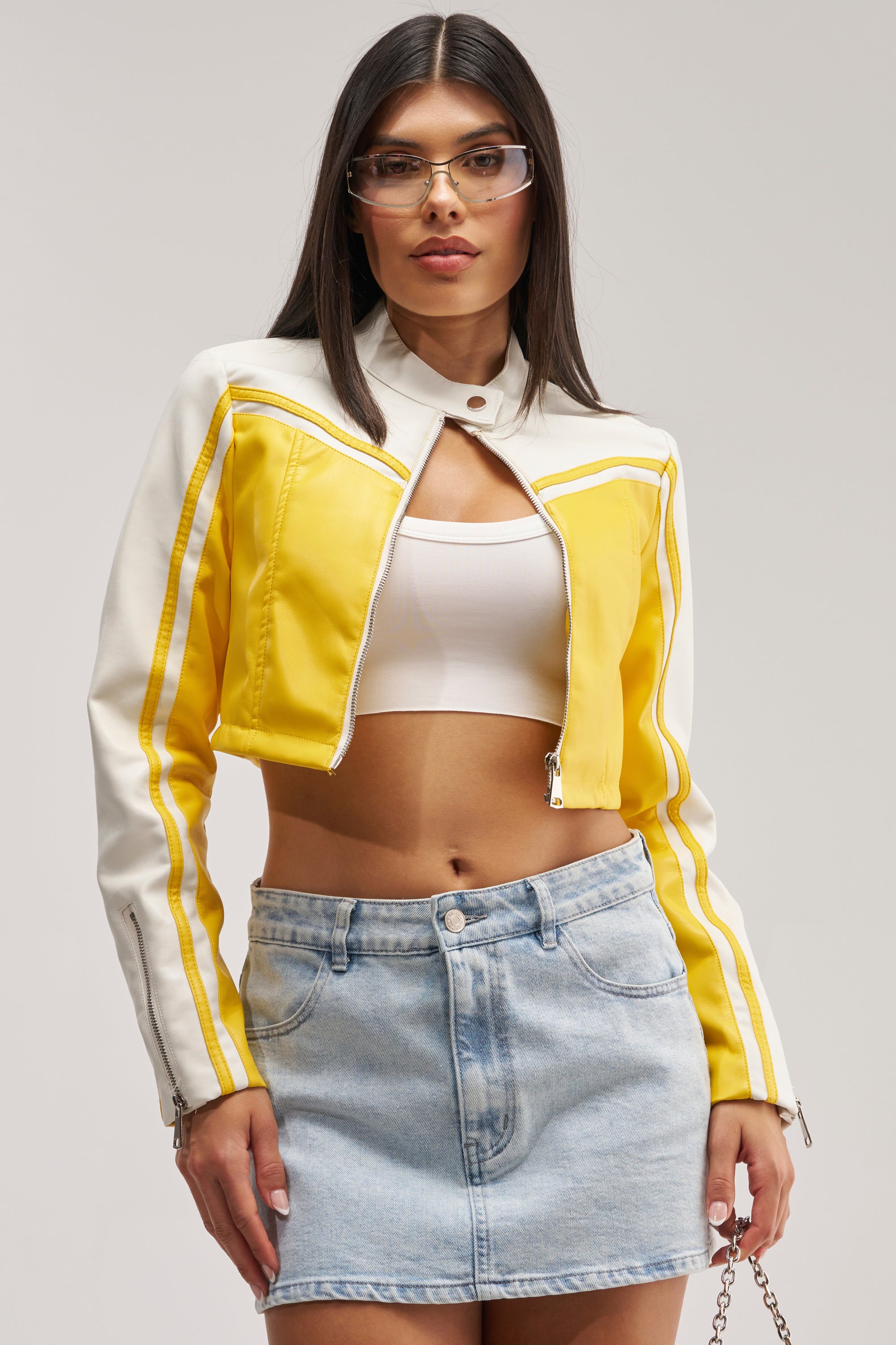 A woman with long dark hair and clear glasses stands against a plain background wearing the WEST LOOP RACER MOTO IN YELLOW, styled with a white crop top and light blue denim mini skirt for an effortlessly cool, streetwear-inspired festival look.