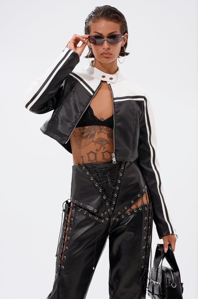 A woman wearing the WEST LOOP RACER MOTO black and white cropped jacket, lace-up leather pants, a visible lace bra, and sunglasses holds a matching bag. She sports wet-look hair and a large torso tattoo.