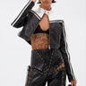 A woman wearing the WEST LOOP RACER MOTO black and white cropped jacket, lace-up leather pants, a visible lace bra, and sunglasses holds a matching bag. She sports wet-look hair and a large torso tattoo.