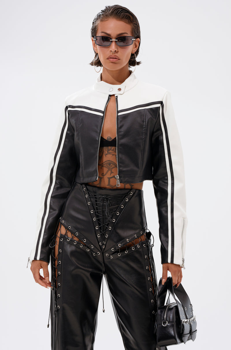 Wearing the WEST LOOP RACER MOTO black and white cropped jacket, high-waisted lace-up black pants with sheer panels, sunglasses, and holding a small black handbag, a woman stands confidently against a plain white background.