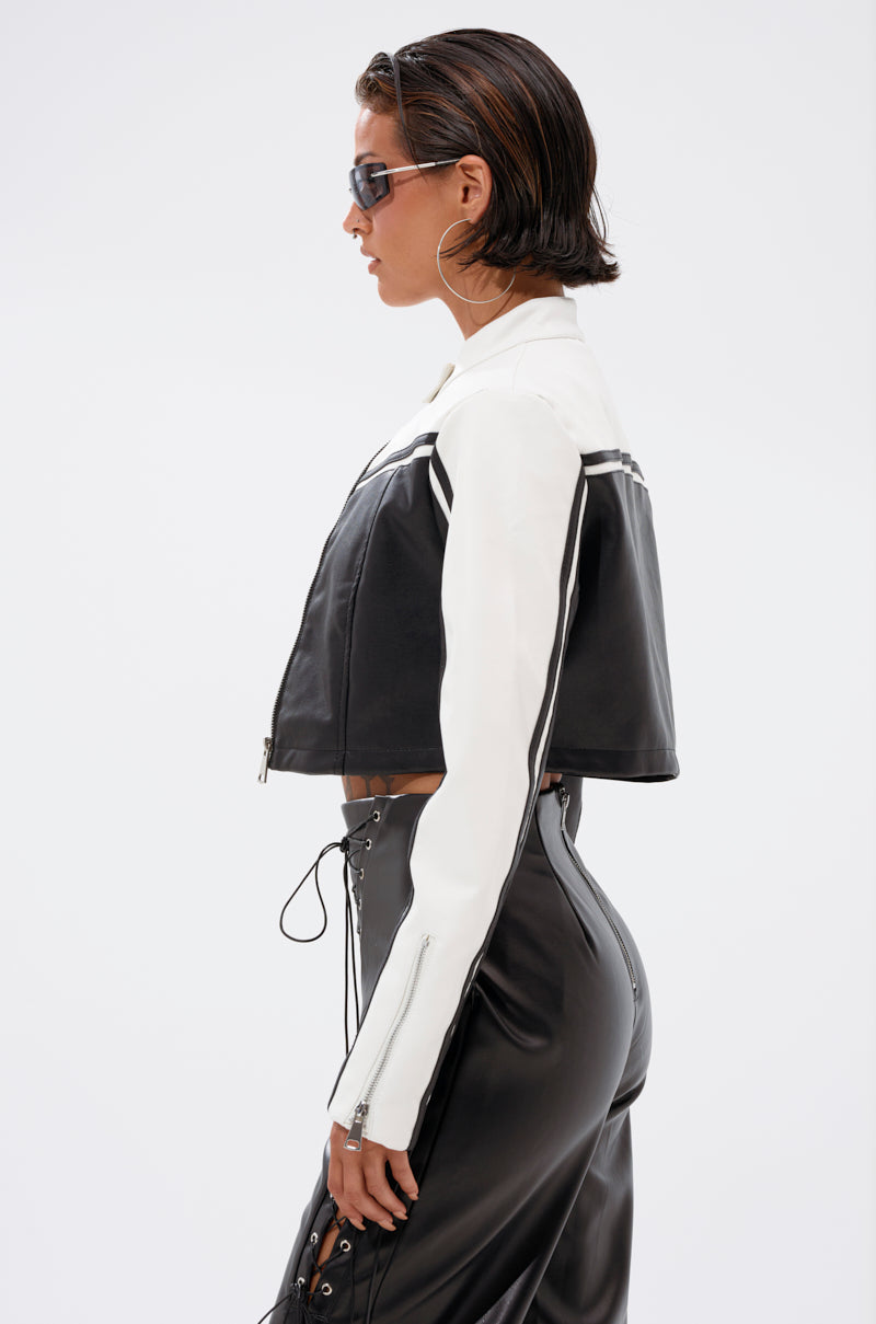 Wearing sunglasses, a person with short, slicked-back hair models the WEST LOOP RACER MOTO—a black and white faux leather cropped moto jacket—paired with high-waisted black lace-up pants, standing in profile against a white background.