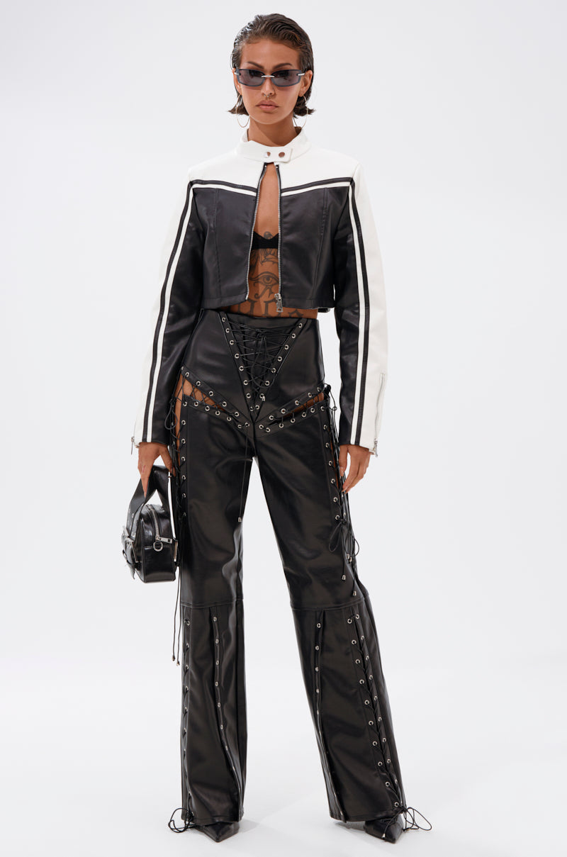 A person stands confidently against a white background wearing the WEST LOOP RACER MOTO black and white cropped jacket, black lace-up cutout pants, sunglasses, and holding a matching bag.