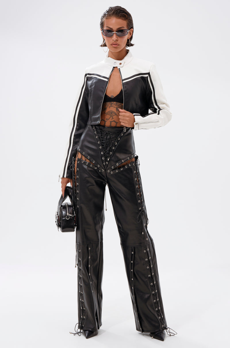 A woman exudes edgy style in the WEST LOOP RACER MOTO black and white faux leather jacket, paired with a sheer patterned top, black lace-up pants, sunglasses, and a small black bag against a plain white background.