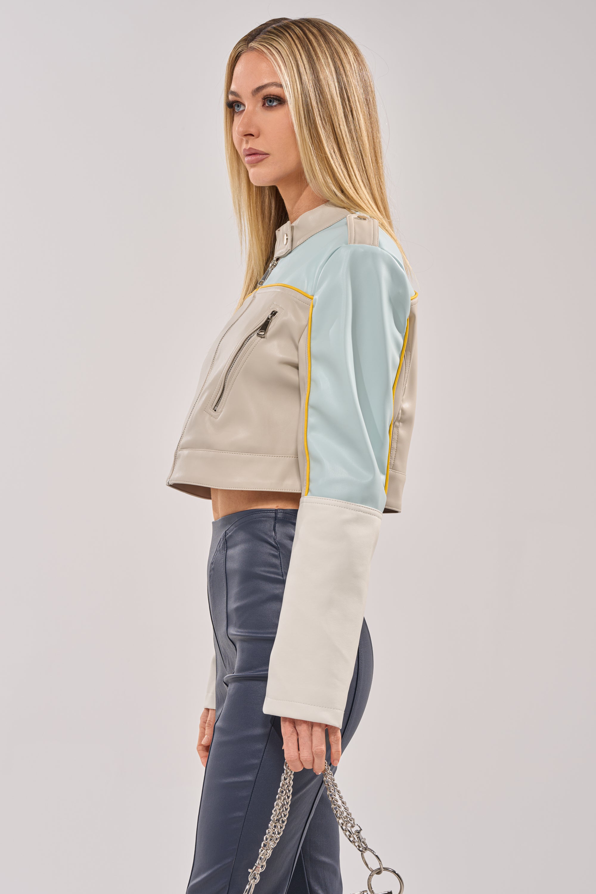 A woman with long, straight blonde hair wears the BETTER LUCK CROPPED MOTO IN LIGHT BLUE, featuring beige, white, and blue colorblocking. She pairs it with high-waisted blue pants and holds silver chains, posing sideways against a plain background. 3
