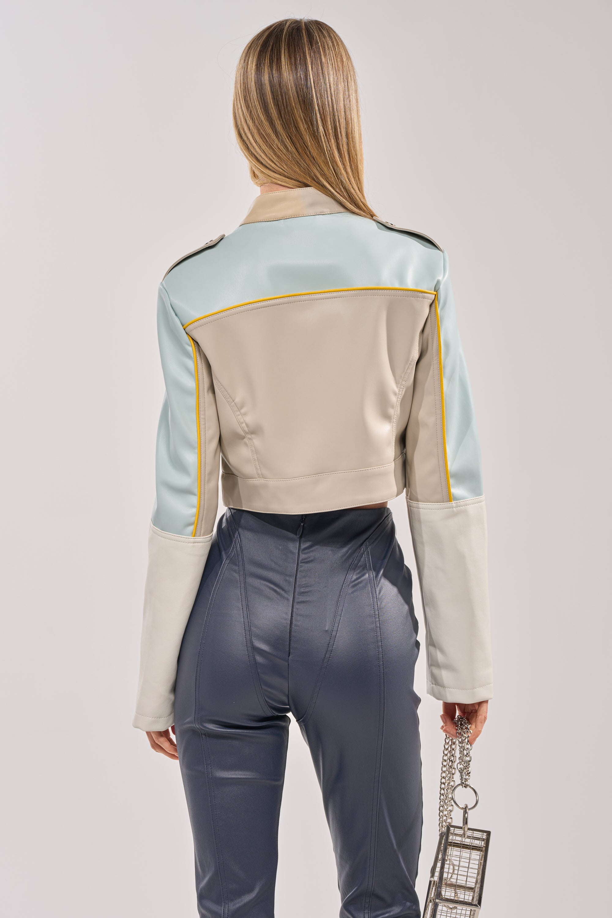 A woman with long, straight hair stands facing away, wearing the BETTER LUCK CROPPED MOTO IN LIGHT BLUE—a colorblocked cropped jacket styled with high-waisted shiny blue pants and a small square handbag for a bold festival look. 4