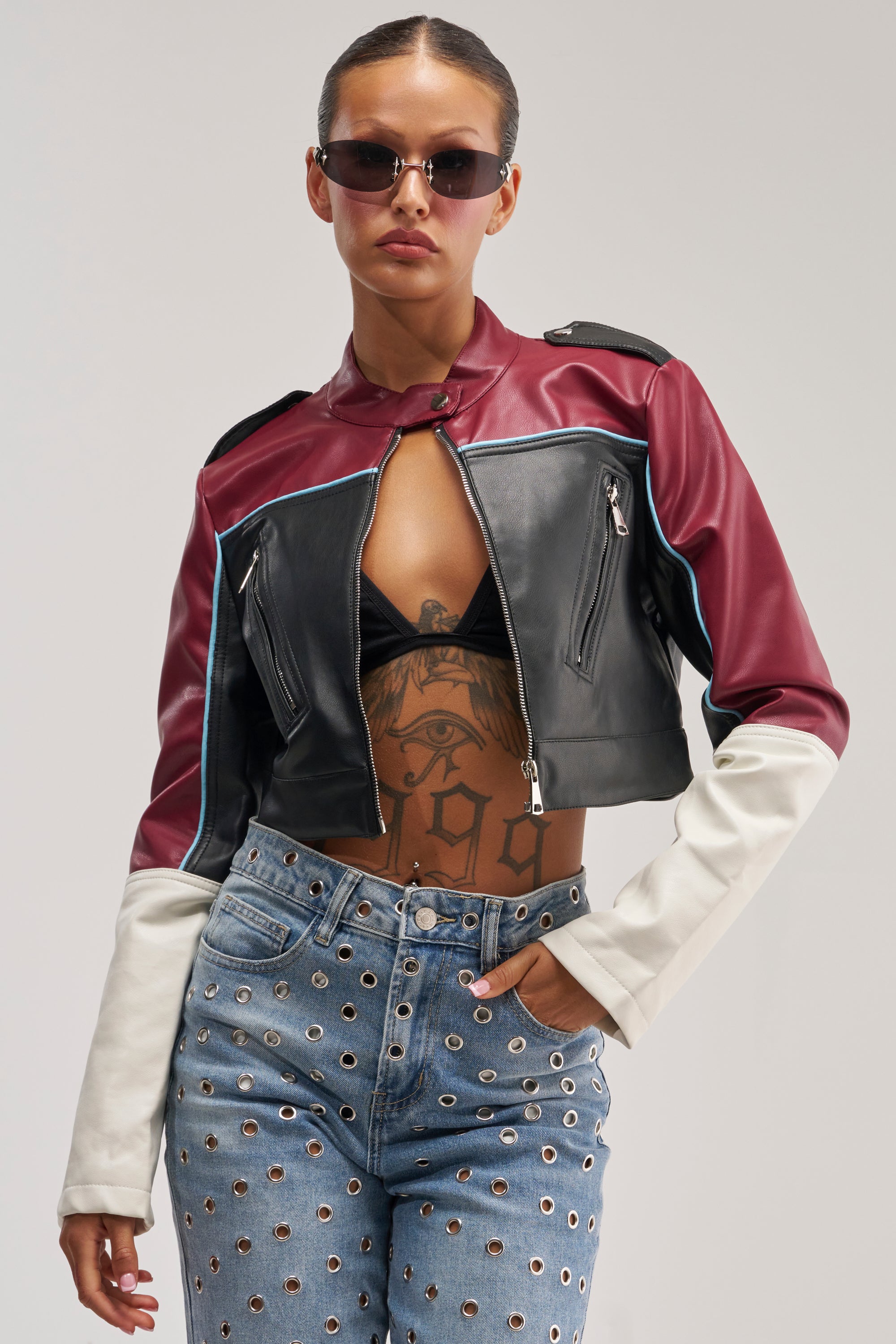 Wearing the BETTER LUCK CROPPED MOTO IN BURGUNDY—a striking maroon, black, and white color block faux leather jacket—she styles it with sunglasses, a black bralette, high-waisted jeans detailed with metal eyelets, and displays her torso tattoos as she confidently poses against a plain background. This standout look is perfect for bold cold-weather layering.