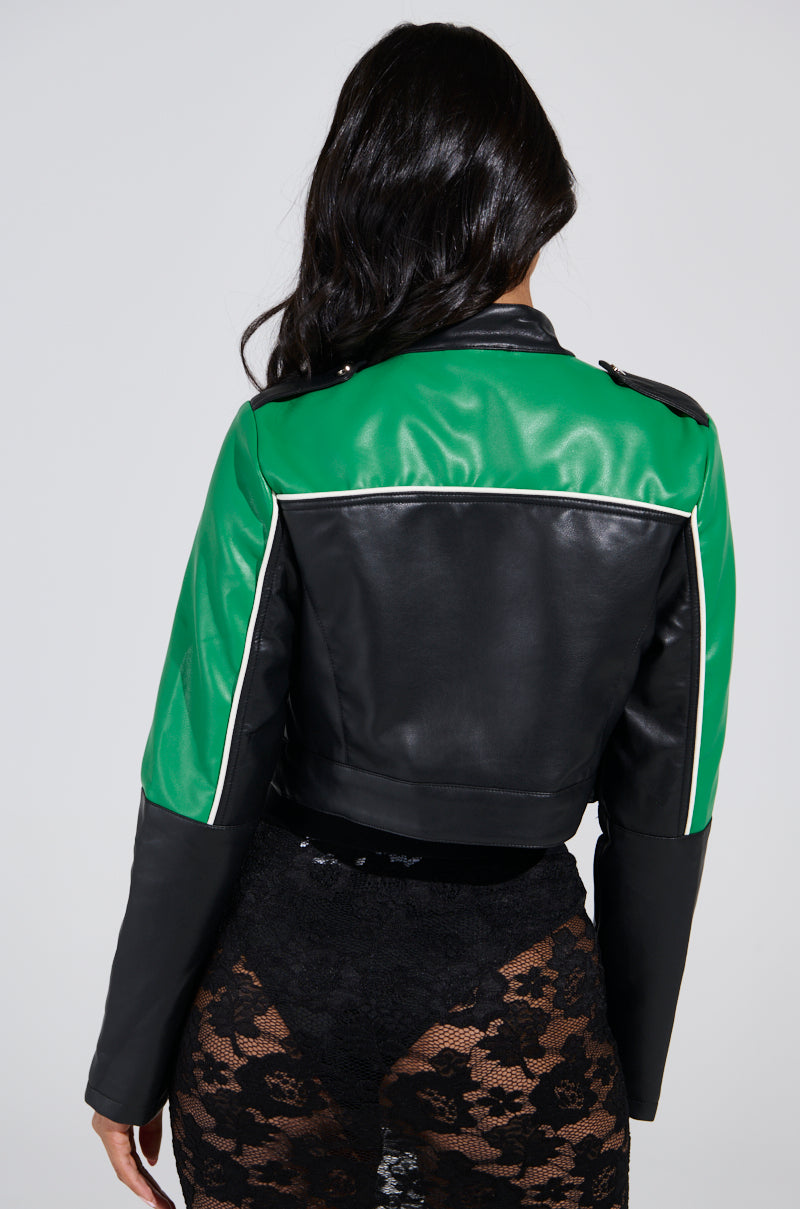 A person with long dark hair, facing away, wears the BETTER LUCK CROPPED MOTO jacket characterized by a green and black color-block design with white trim. They pair it with black lace pants on a plain gray background.
