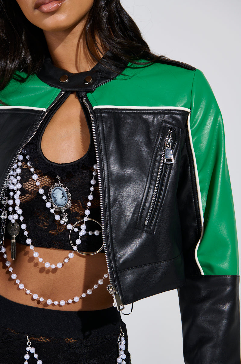 A person is wearing the BETTER LUCK CROPPED MOTO in black and green faux leather, featuring silver zippers, over a black lace top. They accessorize with layered pearl necklaces and a cameo pendant. Their dark, wavy hair partially visible adds to the edgy color-block style.