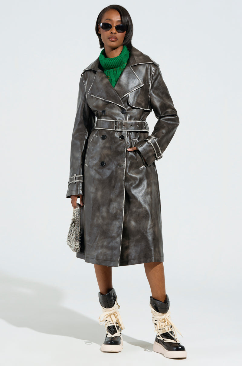 Wearing the ACID RAP FAUX LEATHER TRENCH with acid wash trim, a green sweater, and beige and black boots, the person accessorizes with a silver handbag and dark sunglasses in a studio setting.