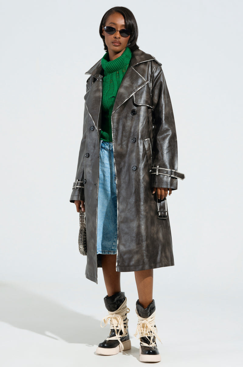 A person sporting the ACID RAP FAUX LEATHER TRENCH, complete with double-breasted button closures, exudes confidence in their sunglasses. They pair it with a green sweater, a blue denim skirt featuring acid wash trim, and lace-up boots while carrying a small handbag.