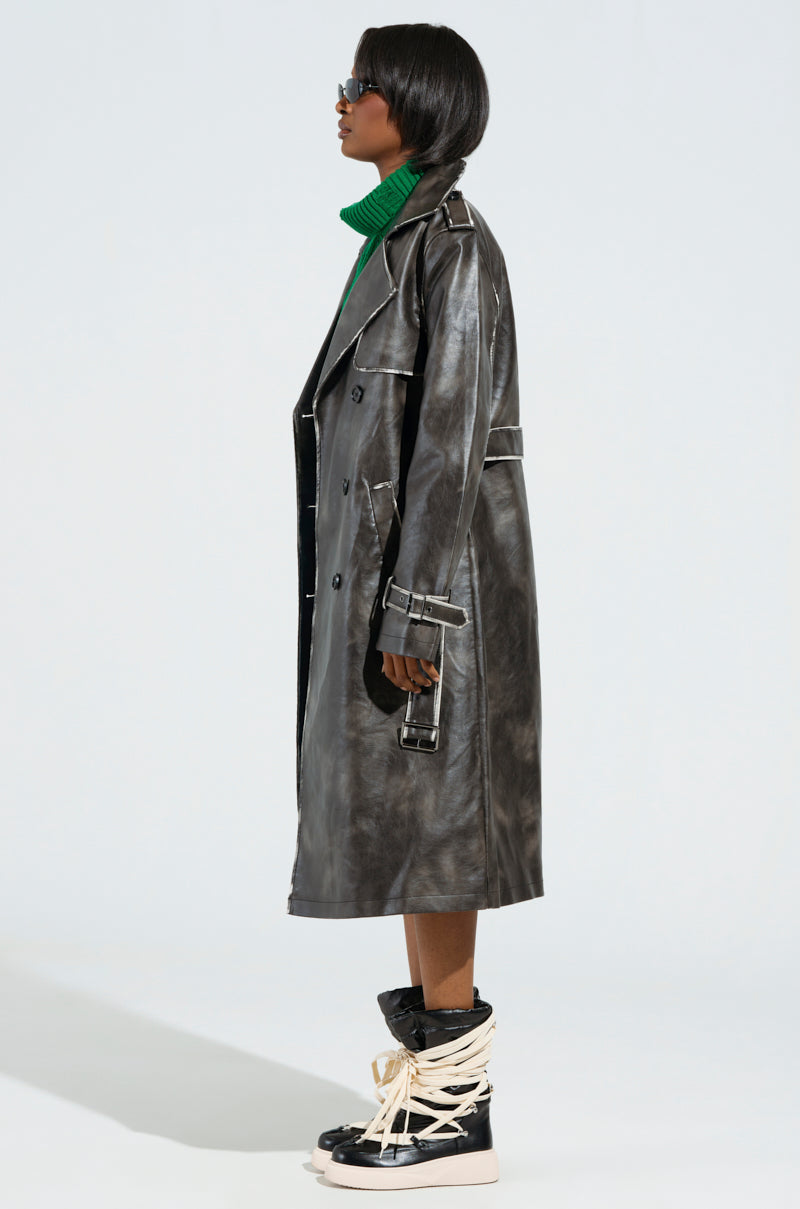 Clad in the ACID RAP FAUX LEATHER TRENCH over a green turtleneck, a person stands in profile. They accessorize with sunglasses and high-top sneakers featuring prominent crisscrossed laces, all set against a plain light background.