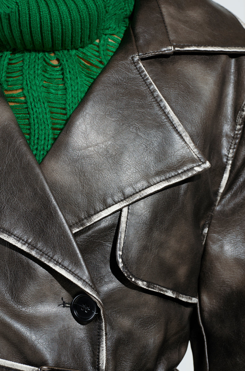 A close-up of an individual wearing the ACID RAP FAUX LEATHER TRENCH, characterized by its dark color with white piping, alongside a dark green turtleneck sweater. The trench features large lapels and double-breasted button closures, highlighting the strikingly visible detailed textures of the materials.